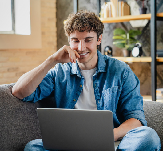 A young man sits on a couch, smiling while looking at a laptop. He is wearing a denim shirt and white t-shirt. There are shelves with books and decor in the background.