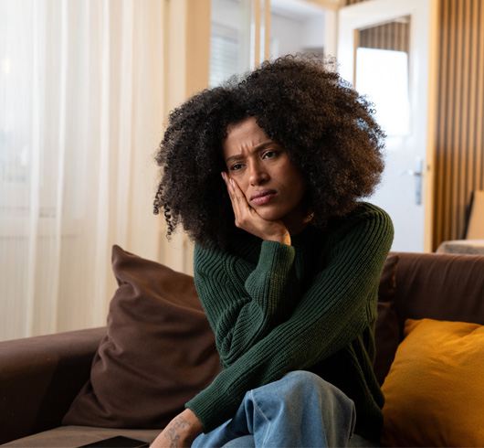 A woman with curly hair sits on a couch, resting her face on her hand, looking concerned or deep in thought in a cozy living room.