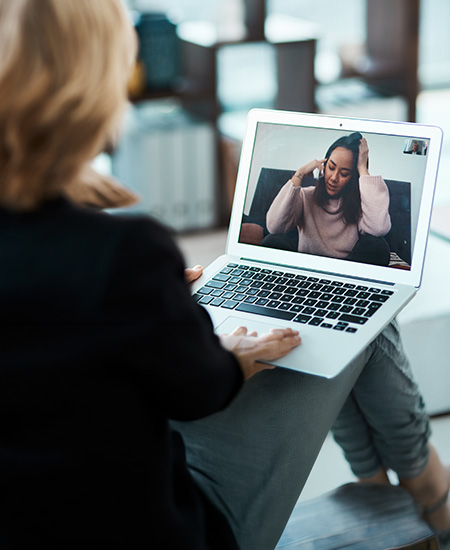 A person sits with a laptop on their lap, having a video call with another person who appears distressed, holding their head with one hand and gesturing while talking.