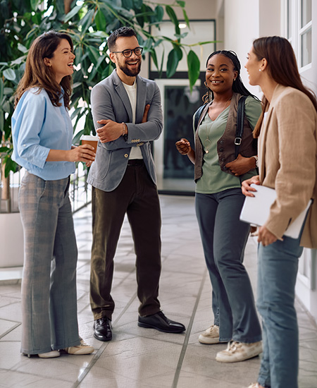 Four people stand together in a hallway having a cheerful conversation. Casually dressed and relaxed, they smile as one holds a coffee cup and another a laptop, amid large plants in the background.