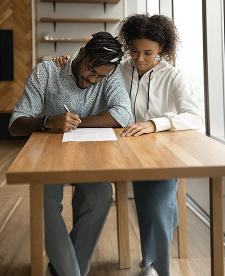 A man sits at a wooden table signing a document while a woman stands beside him, resting her hand on his shoulder supportively. They are indoors near large windows filled with natural light.