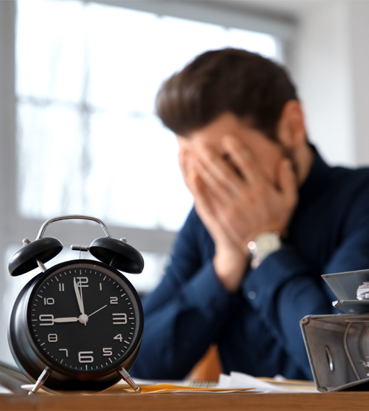 A man sits at a desk with his face in his hands, appearing stressed. A black alarm clock showing 8:20 is in the foreground.