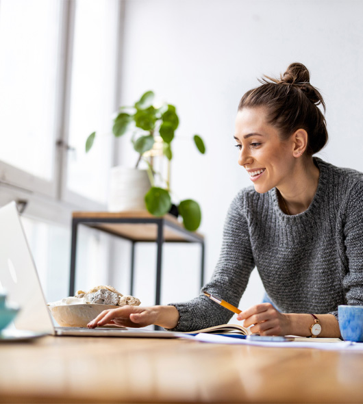 A woman in a grey sweater smiles while working at a laptop, holding a pen. She is seated at a desk with papers and a plant in the background by a window.