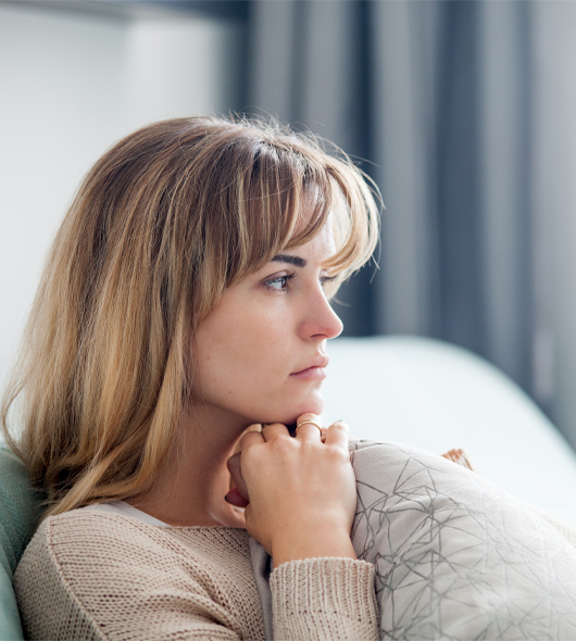A young woman with light brown hair sits on a couch, gazing thoughtfully out of frame, her hands resting under her chin and a patterned pillow in front of her. Soft natural light enters from the side.
