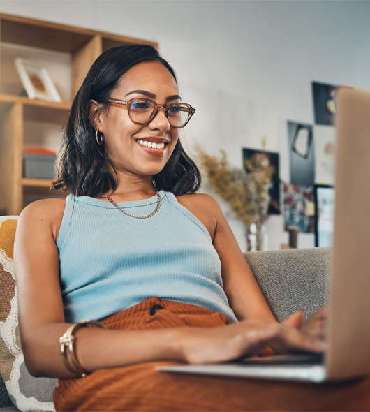 A woman wearing glasses and a blue tank top smiles while using a laptop, sitting on a couch in a cozy living room with shelves and wall art in the background.