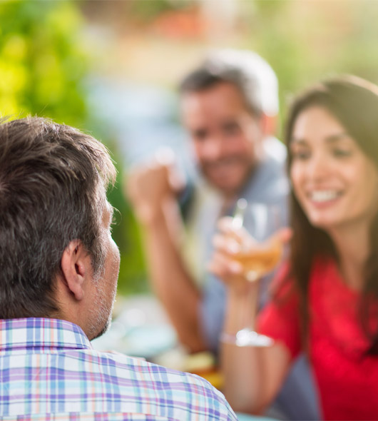 Three adults sit together outdoors, smiling and talking. The man in the foreground faces away from the camera, while the two people in the background appear to be enjoying a conversation and drinks. The setting is bright and casual.