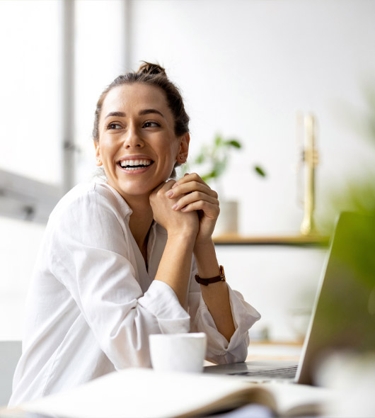 A woman in a white shirt sits at a desk with a laptop and a coffee cup, smiling and looking to the side in a bright, modern office space.