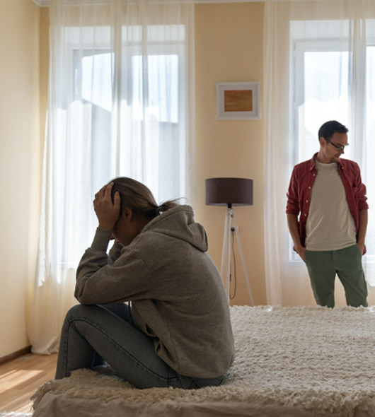 A woman sits on a bed with her head in her hands, looking distressed. A man stands nearby, looking at her with concern. The room is softly lit with natural light from large windows.