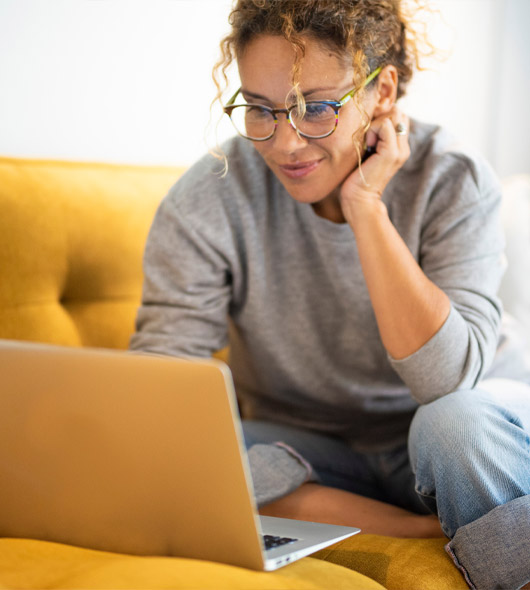 A woman wearing glasses and a gray sweater sits on a yellow couch, smiling as she looks at a laptop on her lap.