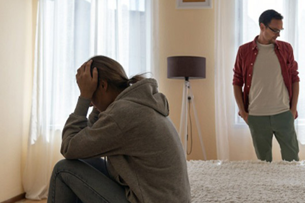 A person sits on a bed with their head in their hands, appearing upset, while another person stands nearby looking away in a softly lit room.