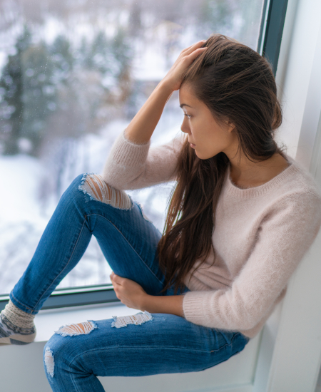 A woman with long hair sits by a window, wearing a light sweater and ripped jeans, looking outside at a snowy landscape with a thoughtful expression.