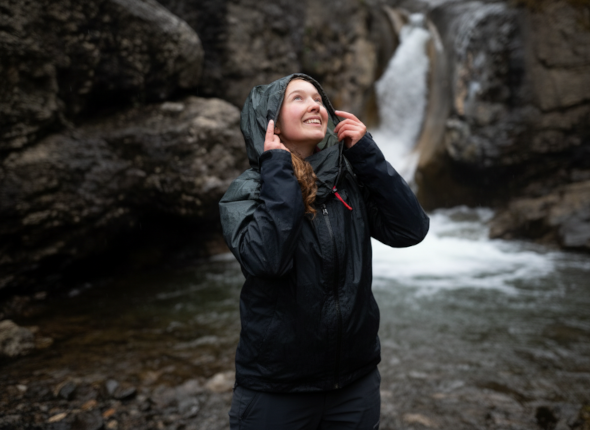 A person in a rain jacket stands near a waterfall, smiling and looking up while holding their hood. Rocky cliffs and water surround them.