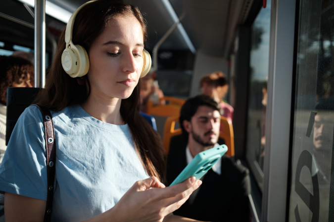 A young woman wearing large headphones looks at her phone while riding a bus. Sunlight streams through the window, and several other passengers are seated in the background.