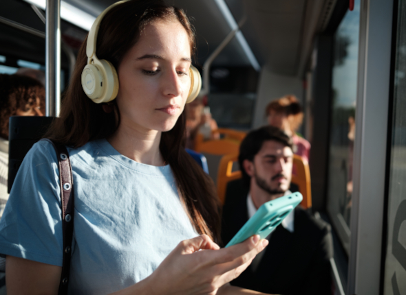 Young woman wearing headphones and a light blue shirt looks at her smartphone while riding a bus. Sunlight comes through the window, and other passengers sit nearby.