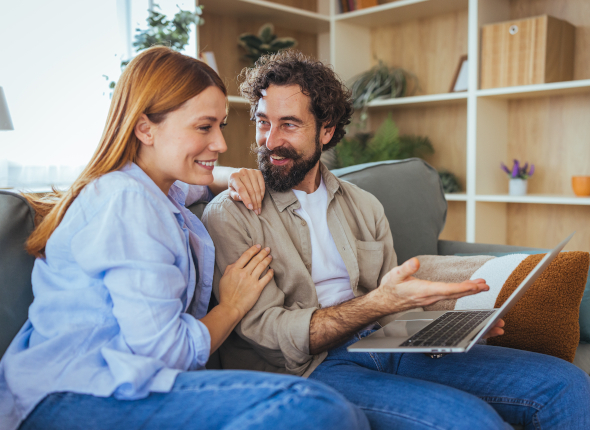 A smiling couple sits together on a couch, looking at a laptop. The man gestures toward the screen while the woman leans in, both appearing happy and engaged in conversation.