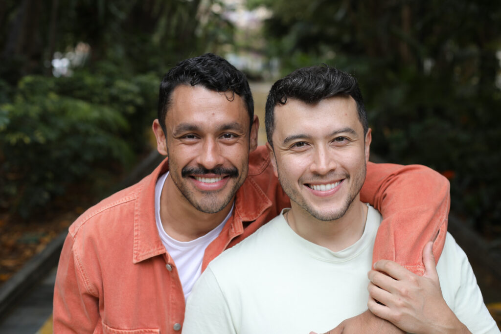 Two men smiling outdoors, one in an orange jacket with his arm around the other, who is wearing a light-colored shirt. Greenery and a path are visible in the blurred background.