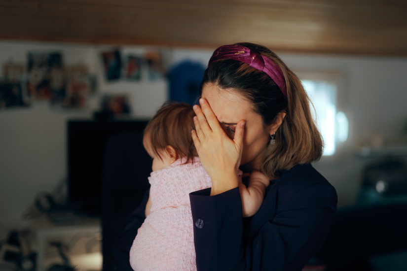 A woman holding a baby covers her face with her hand, appearing stressed or upset, in a dimly lit room with blurred background details.