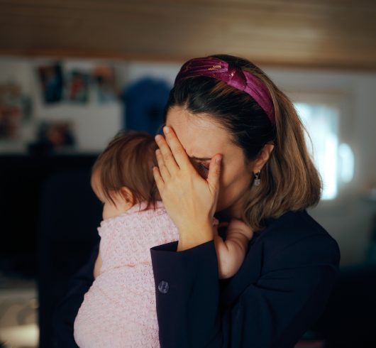 A woman holds a baby while covering her face with one hand, appearing stressed or overwhelmed. The baby is dressed in light pink clothing and rests against her shoulder in a softly lit indoor setting.