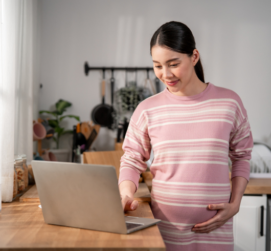 A pregnant woman in a pink striped sweater stands in a kitchen, smiling while using a laptop on the counter and gently touching her belly.