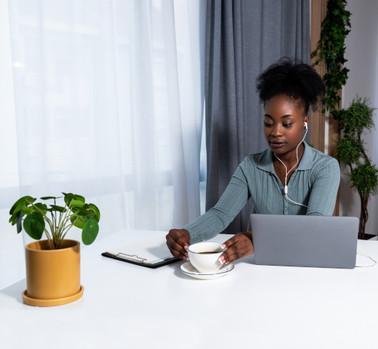 A woman sits at a desk with a laptop, wearing earphones, holding a cup of coffee. A clipboard and a potted plant are on the white table. She appears focused, with light from a window beside her.