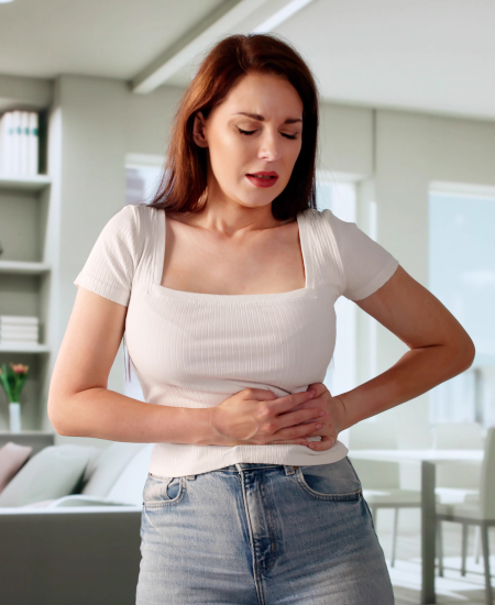 A woman in a white shirt stands indoors, holding her stomach and appearing to be in pain or discomfort.
