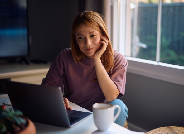 A young woman sits at a desk by a window, looking at a laptop screen with a slight smile. She rests her head on one hand, with a cup of coffee and a small plant nearby.