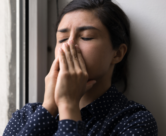 A person with dark hair and a polka-dot shirt stands by a window, eyes closed, pressing their hands to their face as if feeling stressed or overwhelmed.