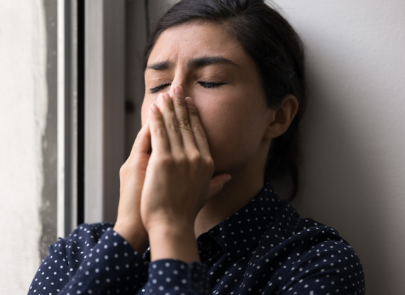 A woman stands by a window with her eyes closed and hands covering her mouth, appearing stressed or upset.