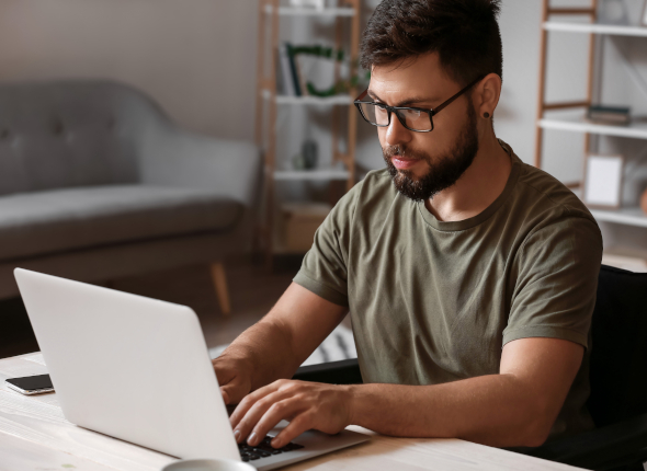 A person with a beard and glasses types on a laptop at a desk in a home office, with shelves and a sofa in the background.