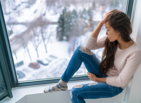 A woman in a sweater and ripped jeans sits by a large window, looking thoughtfully outside at a snowy landscape with trees and parked cars below.