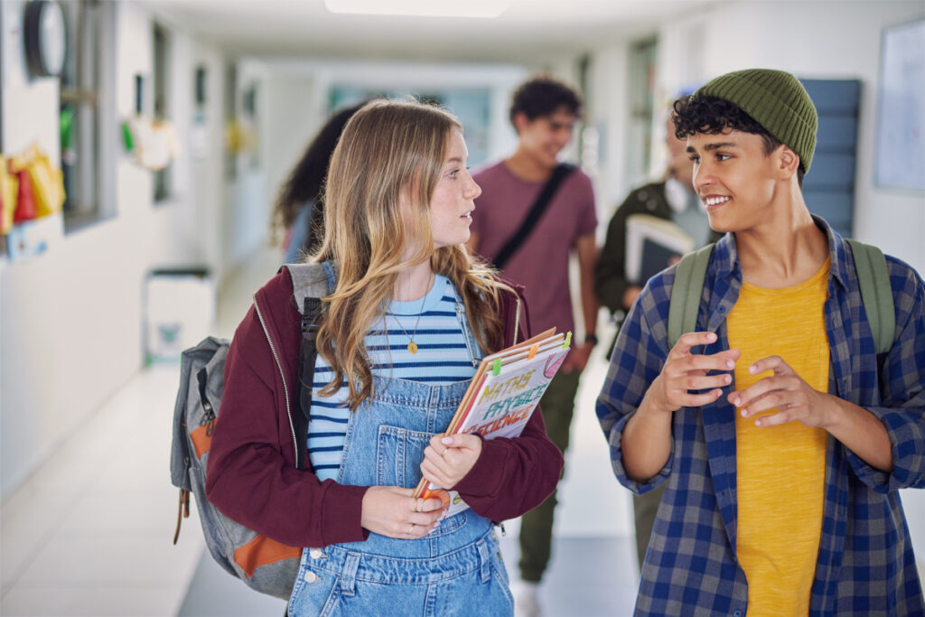 Two teenagers are walking and talking in a school hallway, carrying books and backpacks. Other students are visible in the background. The mood appears friendly and relaxed.