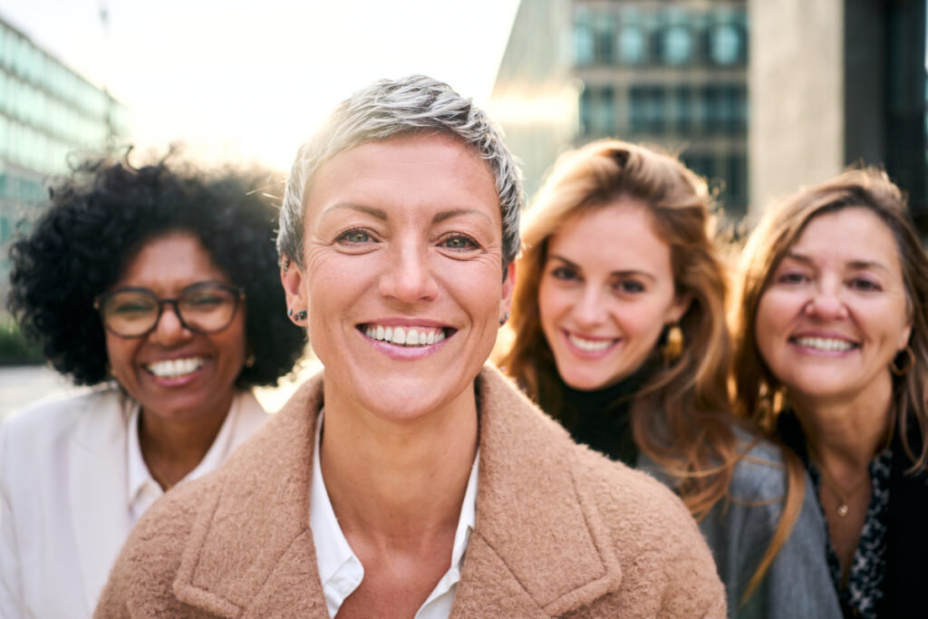 Four women stand close together outdoors, smiling at the camera, with sunlight shining behind them. The focus is on the woman in front, who has short gray hair and is wearing a beige coat.
