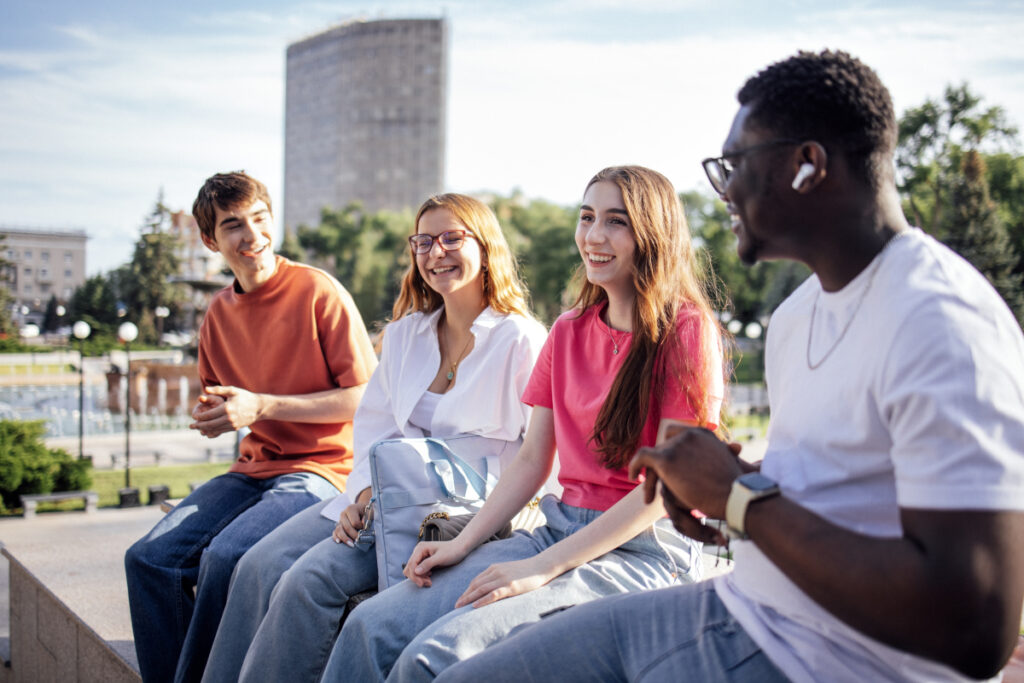 Four young adults sit on a ledge outdoors, smiling and talking on a sunny day. There are trees and a tall building in the background.