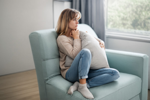 A woman sits on a light blue chair, hugging a pillow to her chest, looking out a window with a thoughtful expression. She wears a beige sweater, blue jeans, and gray socks.