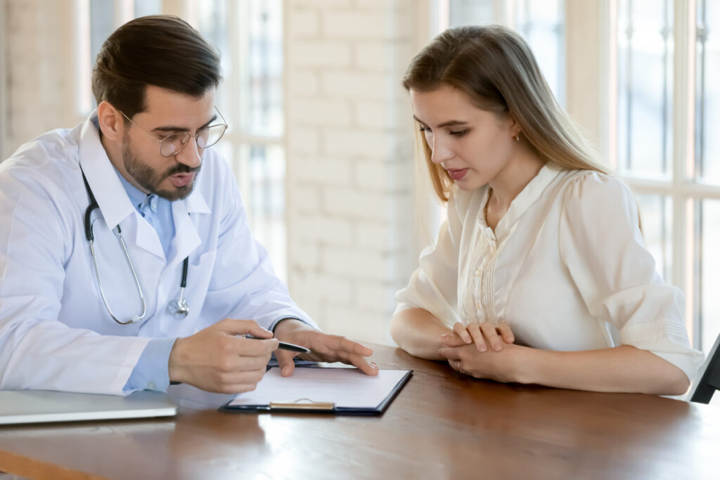 A doctor in a white coat explains information to a woman, showing her a clipboard with documents as they sit together at a wooden table in a well-lit room.